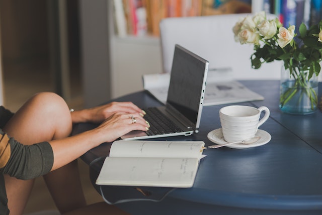 A person using a laptop, with an open notebook and a cup of tea next to it on the table.